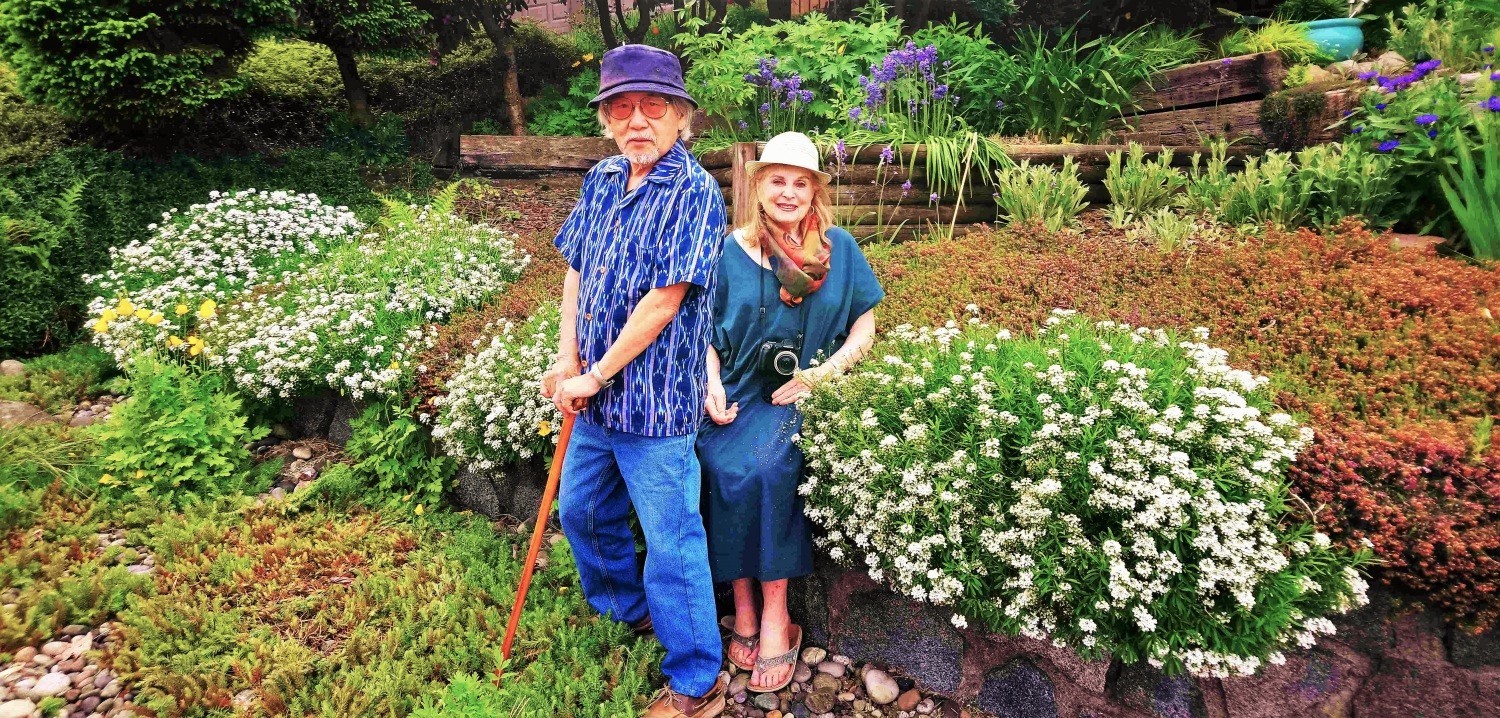 Ken standing in his flowery and terraced front yard with his partner Lorraine